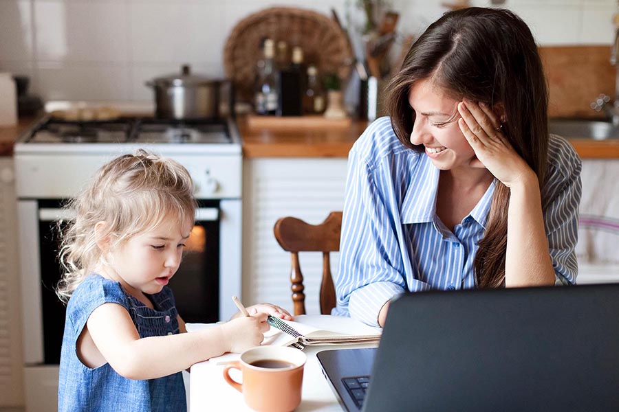 About Our Agency - Mother Uses Her Laptop at the Kitchen Table, Smiling as Her Toddler Daughter Scribbles in a Notebook, With a Cup of Coffee Nearby